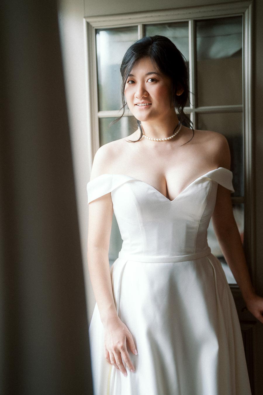 Bride-to-be in an off-the-shoulder white dress standing by a window.