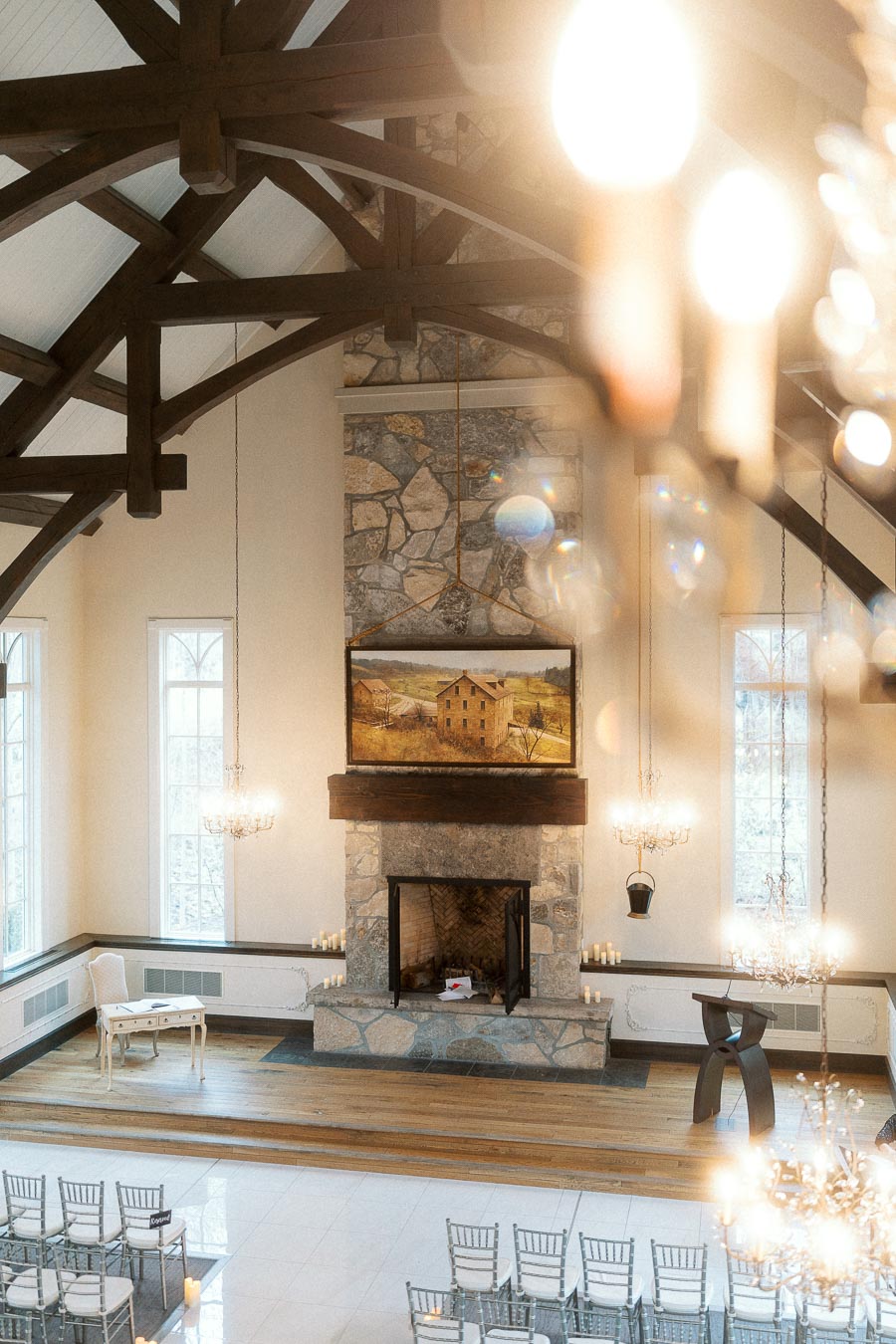 Elegant hall with stone fireplace, exposed wood beams, chandeliers, and ceremony chairs