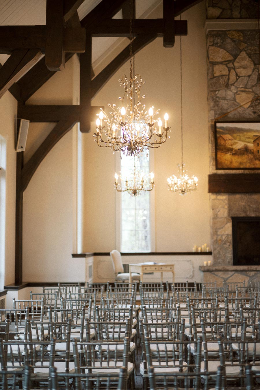 Rows of ceremony chairs beneath chandeliers in a rustic indoor venue