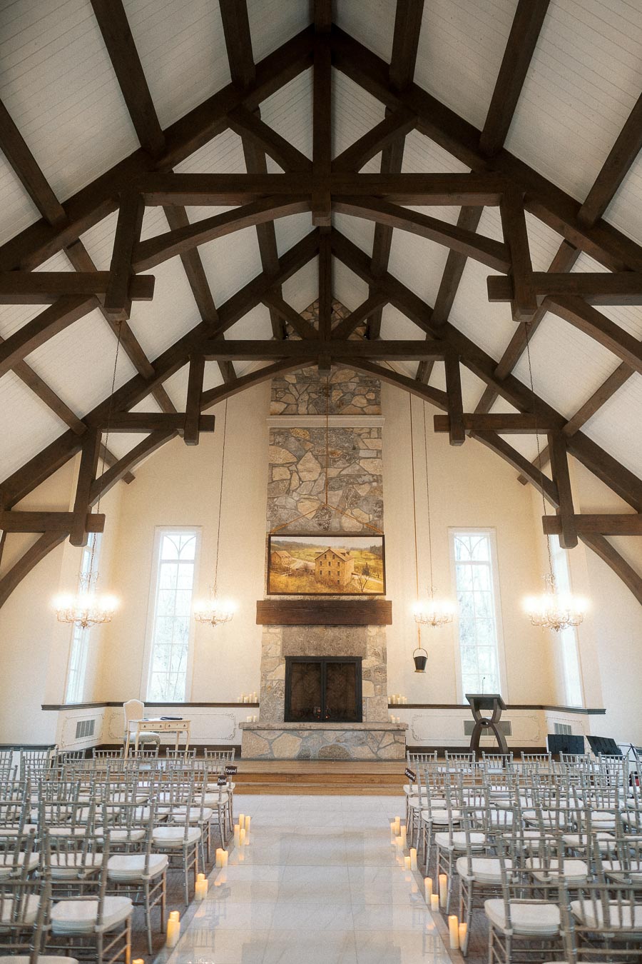 Indoor ceremony space with exposed wood beams, stone fireplace, chairs, and aisle candles