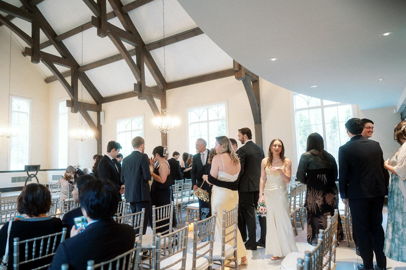 Wedding guests mingling between ceremony chairs in a bright hall with exposed wooden beams.