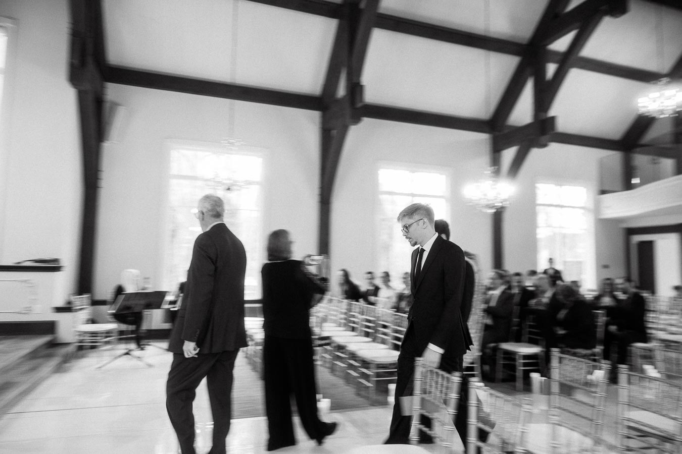 People in formal attire walk through a bright ceremony room with seated guests.