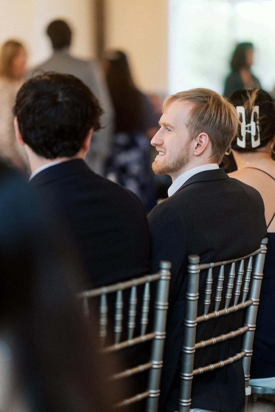 Man in a suit smiling while seated among guests at an indoor event