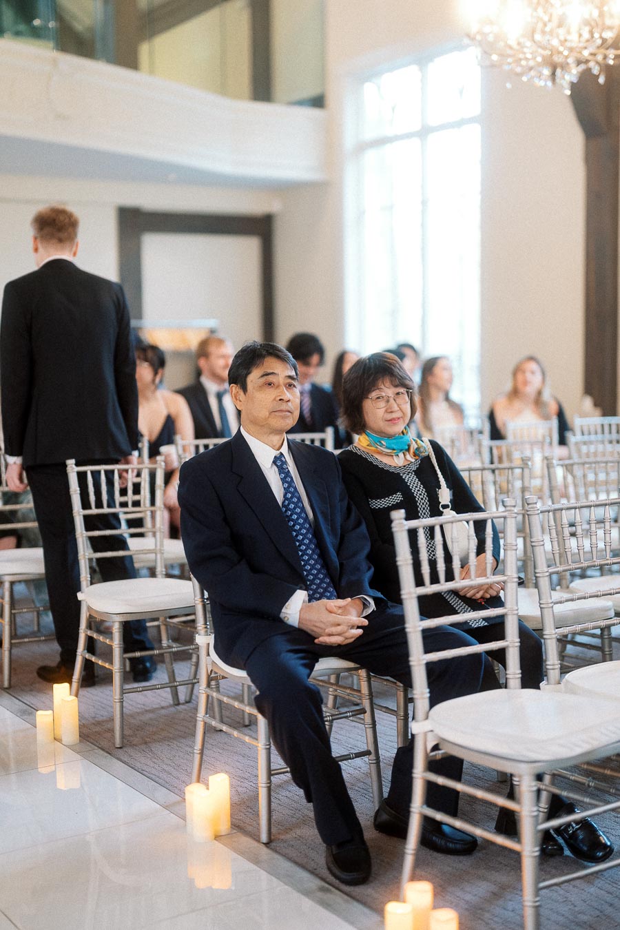 Guests seated in silver chairs during an indoor ceremony with candles and chandelier light