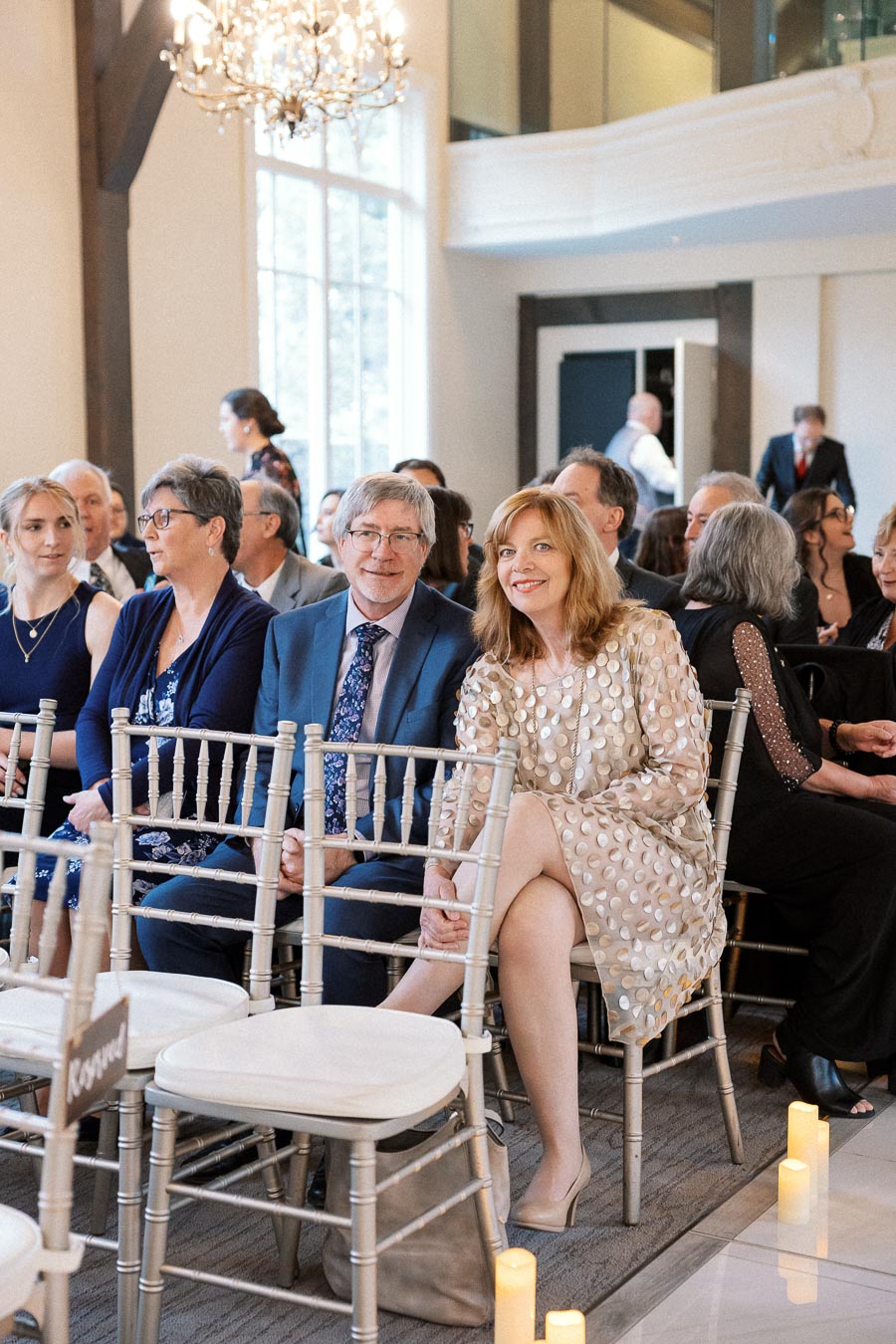 Guests seated in a bright ceremony room with chandeliers and silver chairs.