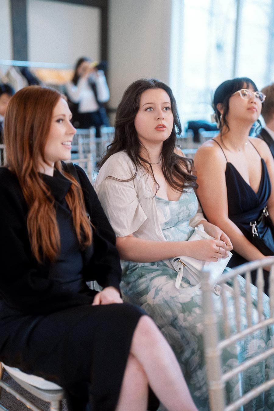 Three women seated in chairs, watching an indoor event attentively.