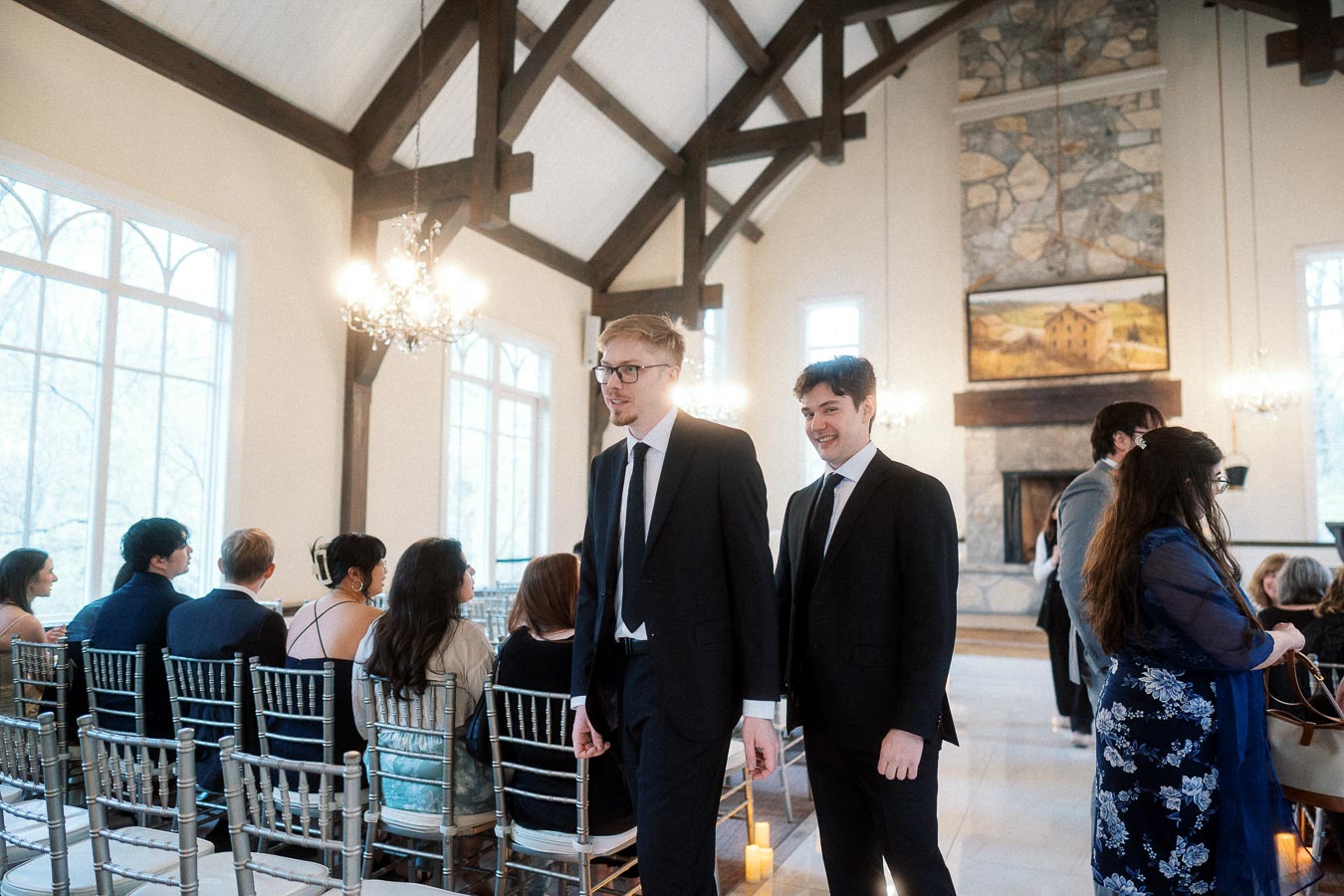 Two men in black suits walk down an aisle in a bright ceremony hall.
