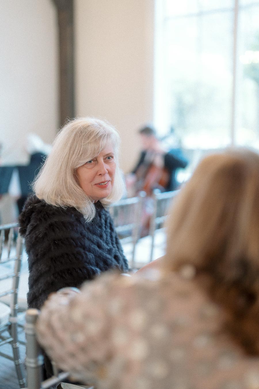Woman with blonde hair seated indoors, speaking with another guest at a formal event.
