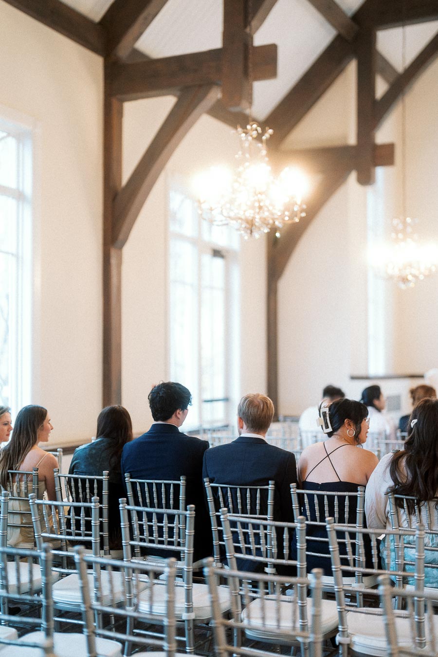 Guests seated in silver chairs inside a bright hall with timber beams and chandeliers
