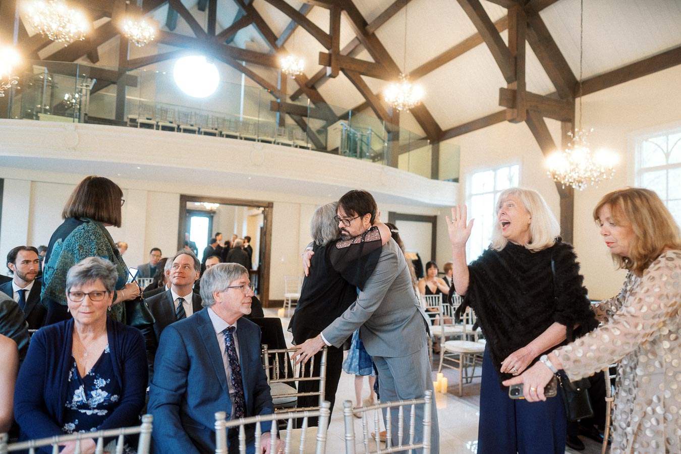 Guests greet and hug inside an elegant hall with chandeliers and wooden beams.