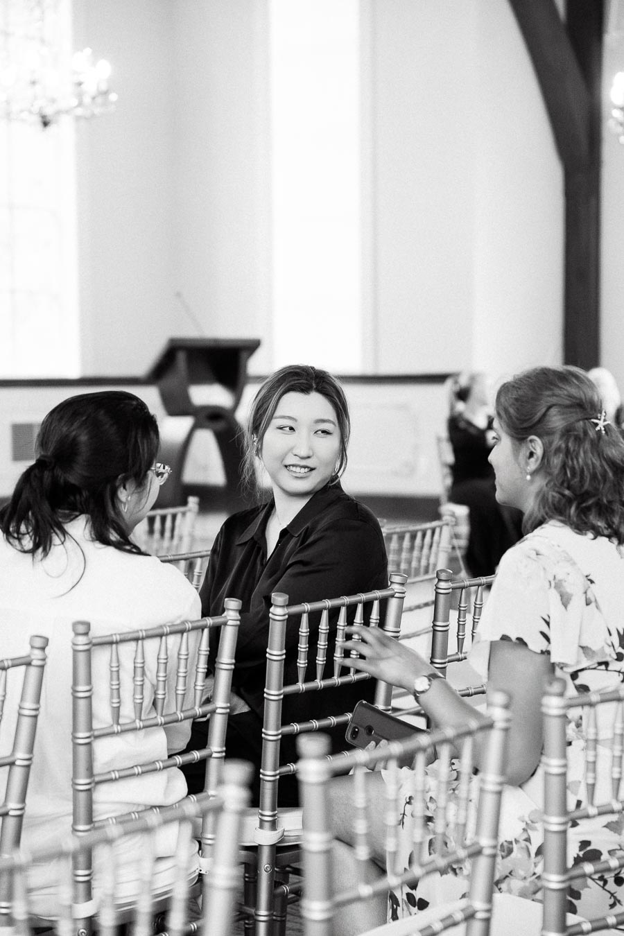 Three women seated among event chairs, talking in a bright indoor venue.