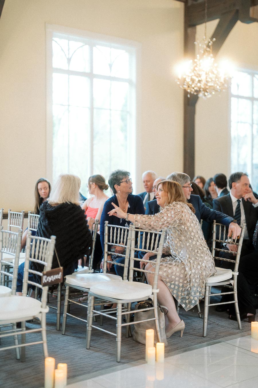 Guests seated on silver chairs talking before an indoor ceremony.