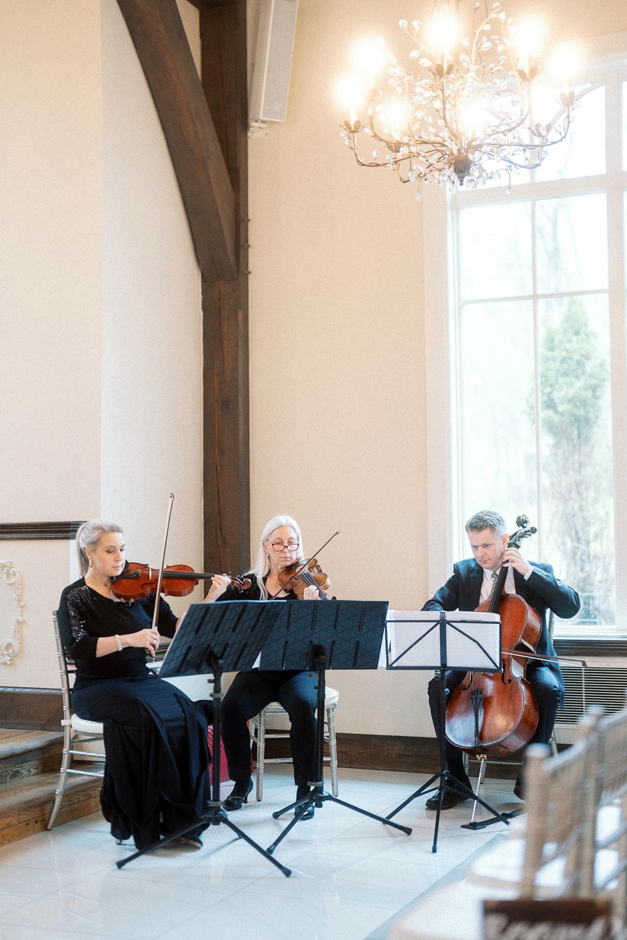 String trio performing indoors beneath a chandelier near a tall window