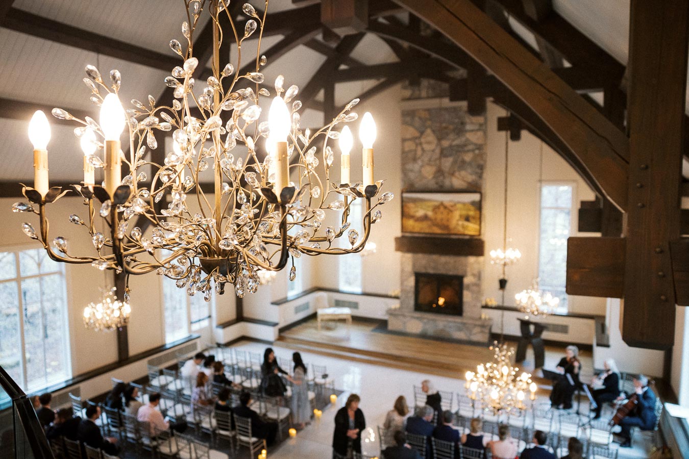 Ornate chandelier overlooking guests seated in a vaulted event hall with a stone fireplace