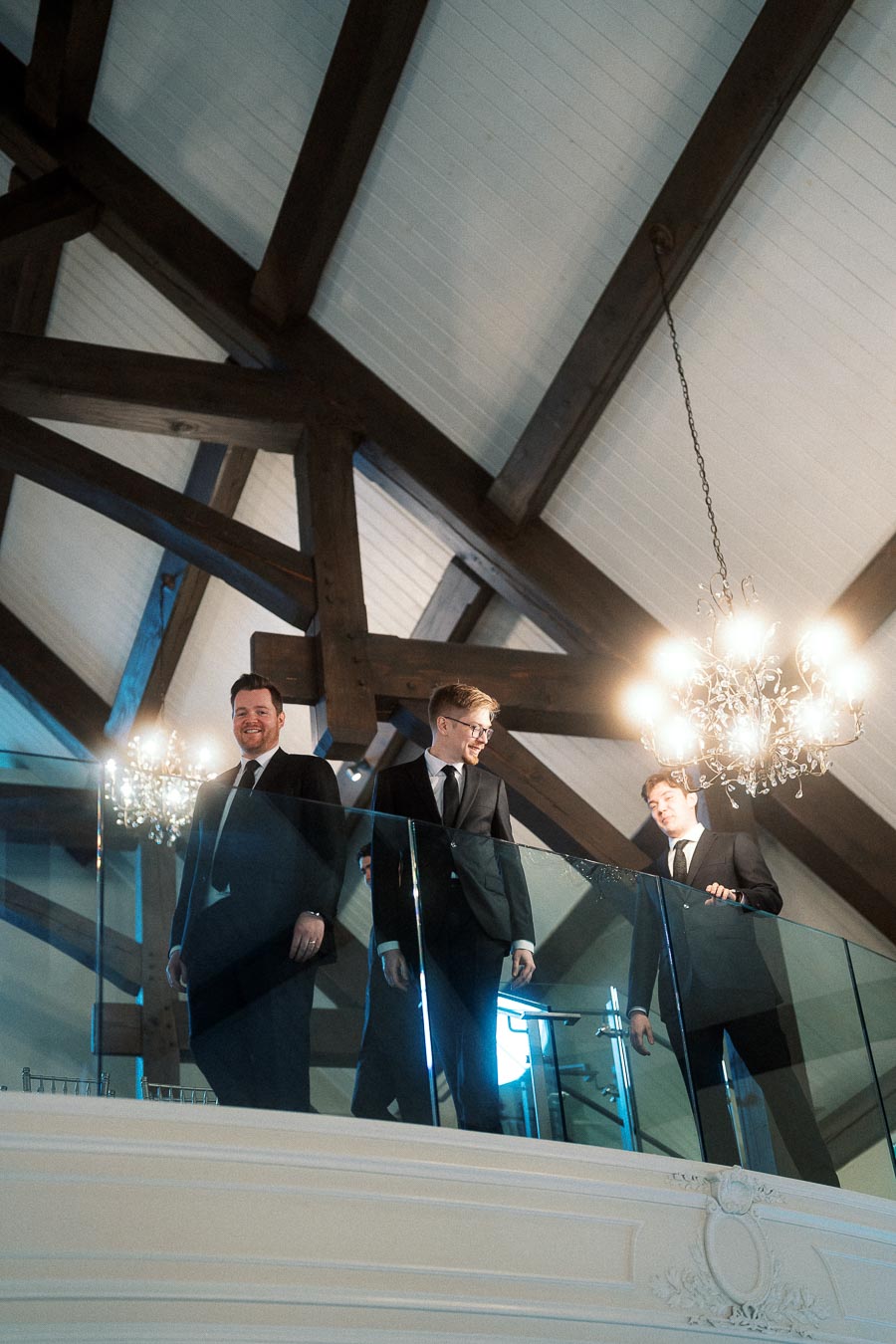 Three men in suits standing on an indoor balcony beneath chandeliers and wooden beams