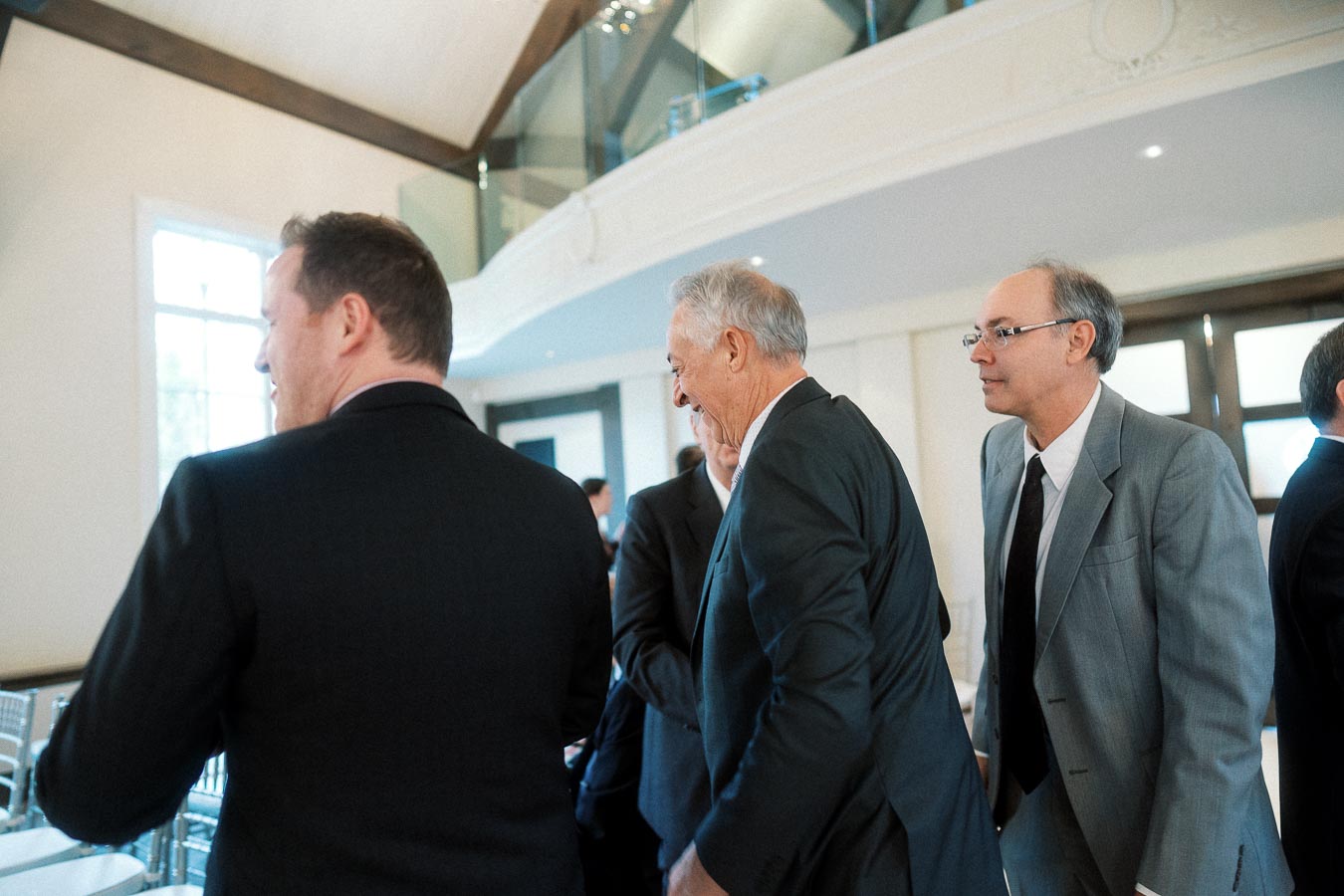 Men in suits talking inside a bright event space with a balcony.