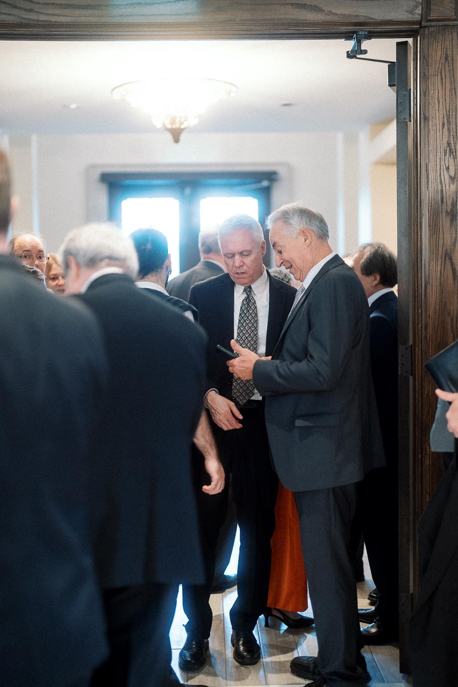 Guests in formal suits gather indoors while two men look at a phone.