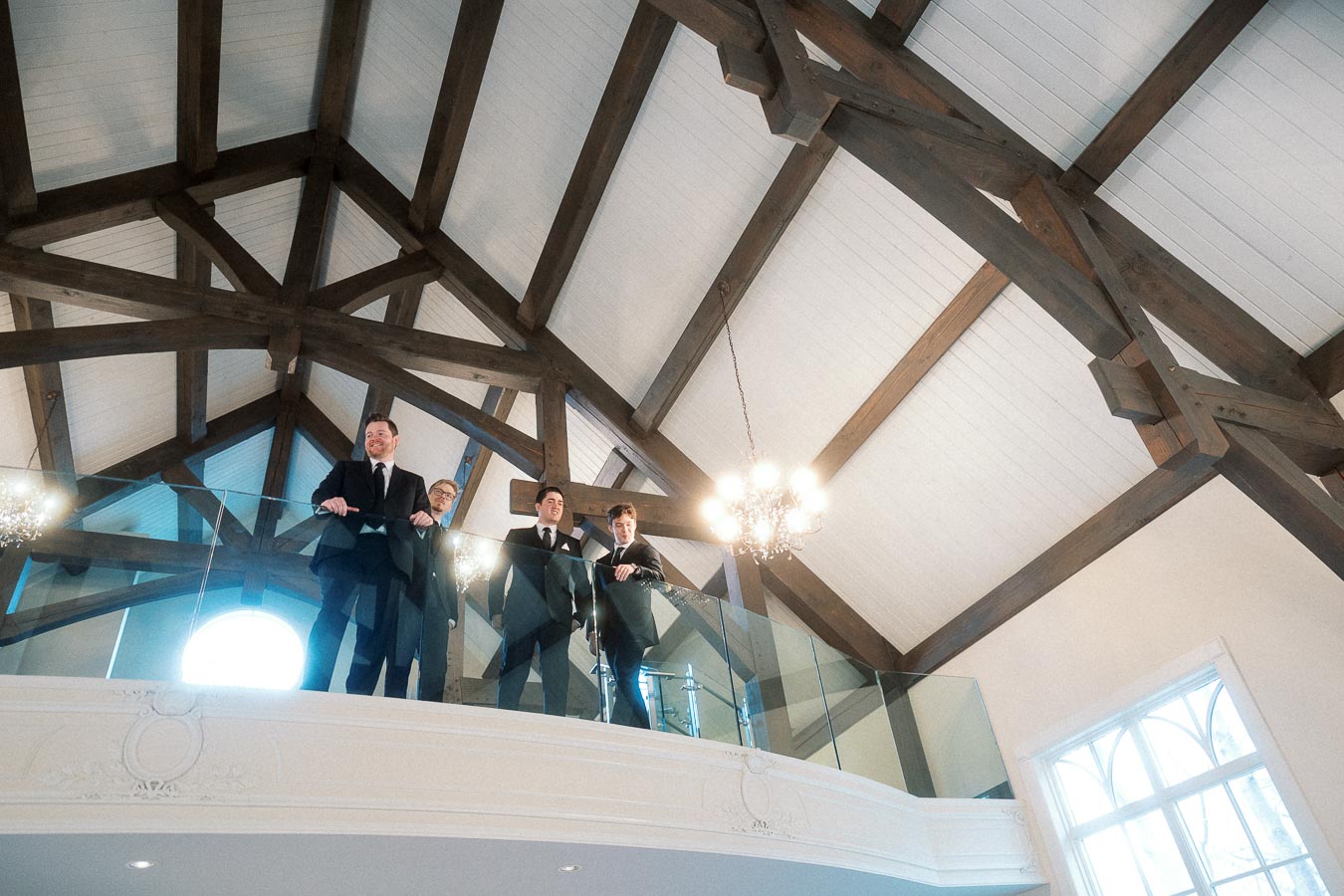 Four men in suits standing on an indoor balcony beneath exposed wooden beams
