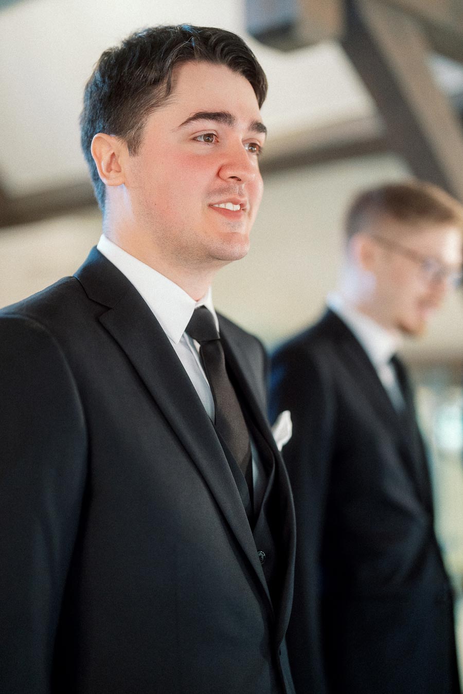Man in a black suit standing indoors with another suited man in the background