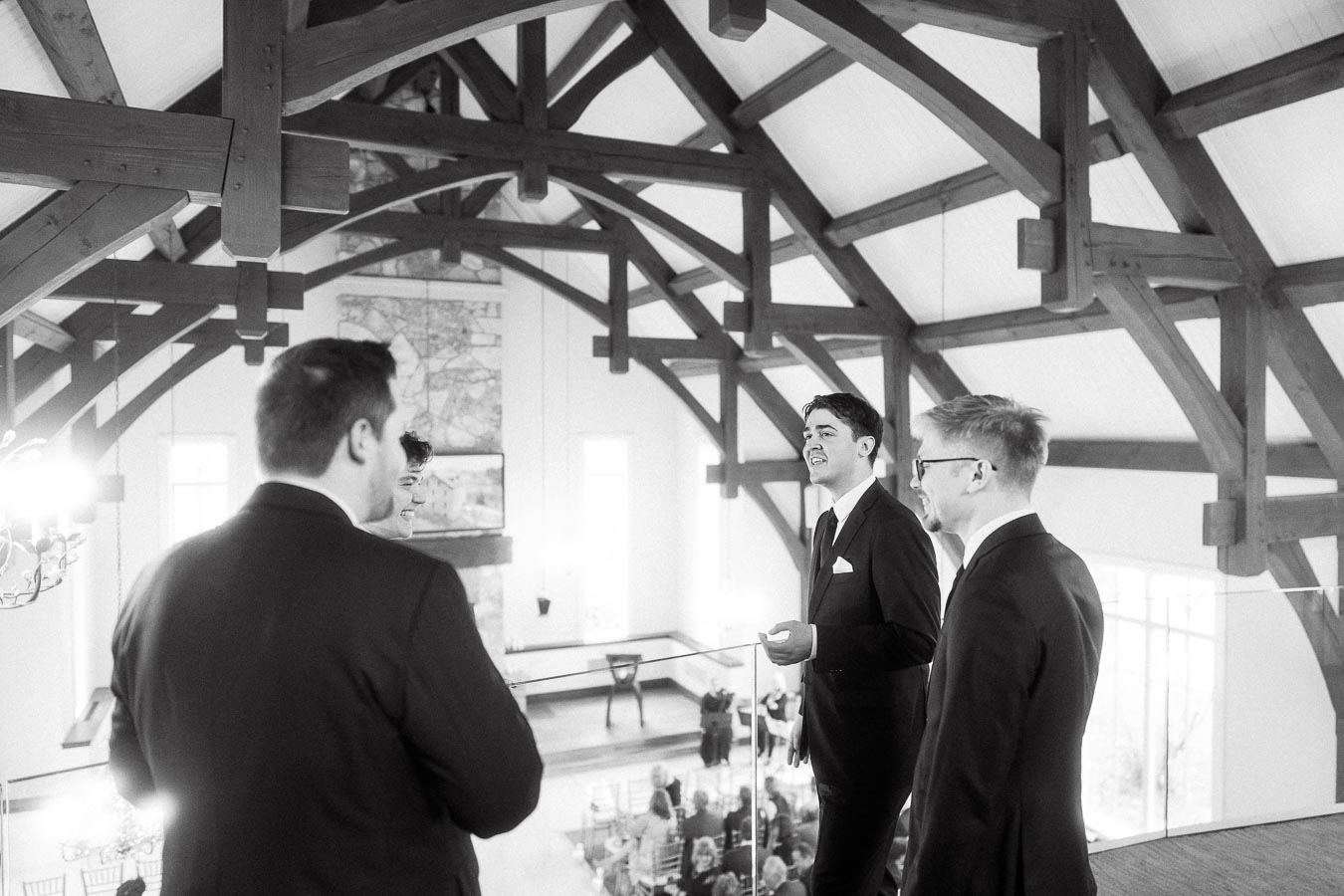 Men in suits talking and laughing on a balcony beneath exposed wooden beams