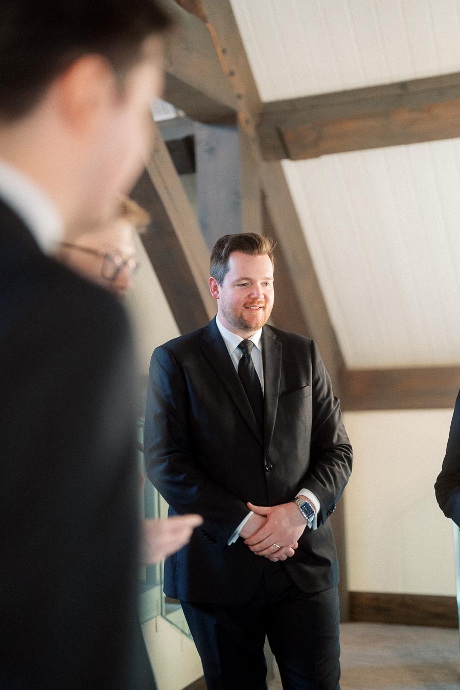 Man in a black suit standing indoors near exposed wooden beams