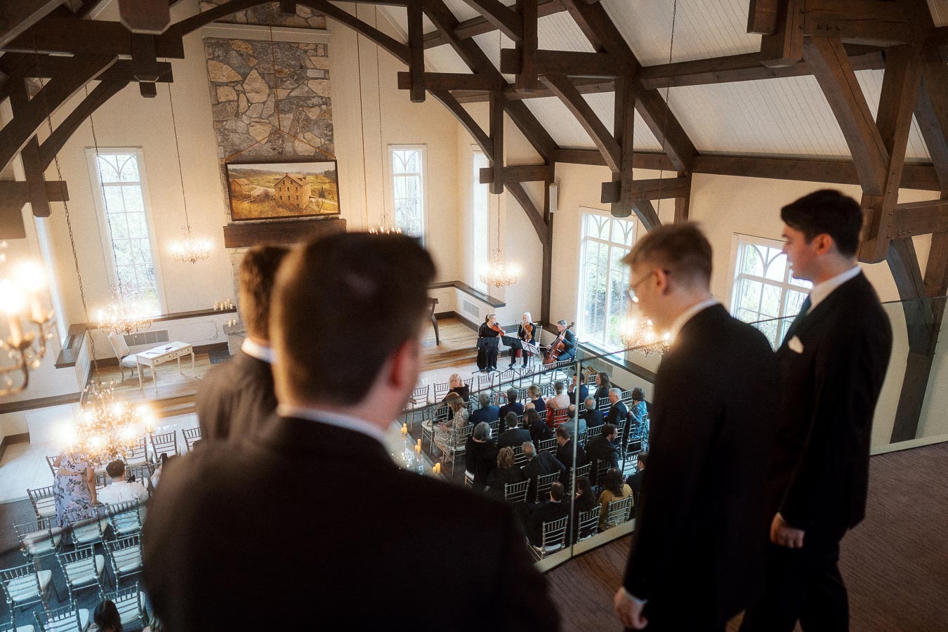 Men in suits overlook an indoor ceremony from a balcony in a timber-framed hall.