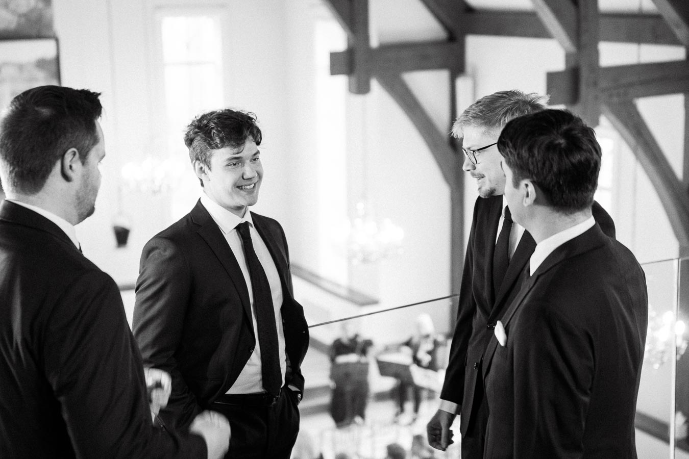 Four men in suits talking indoors beside exposed wooden beams.