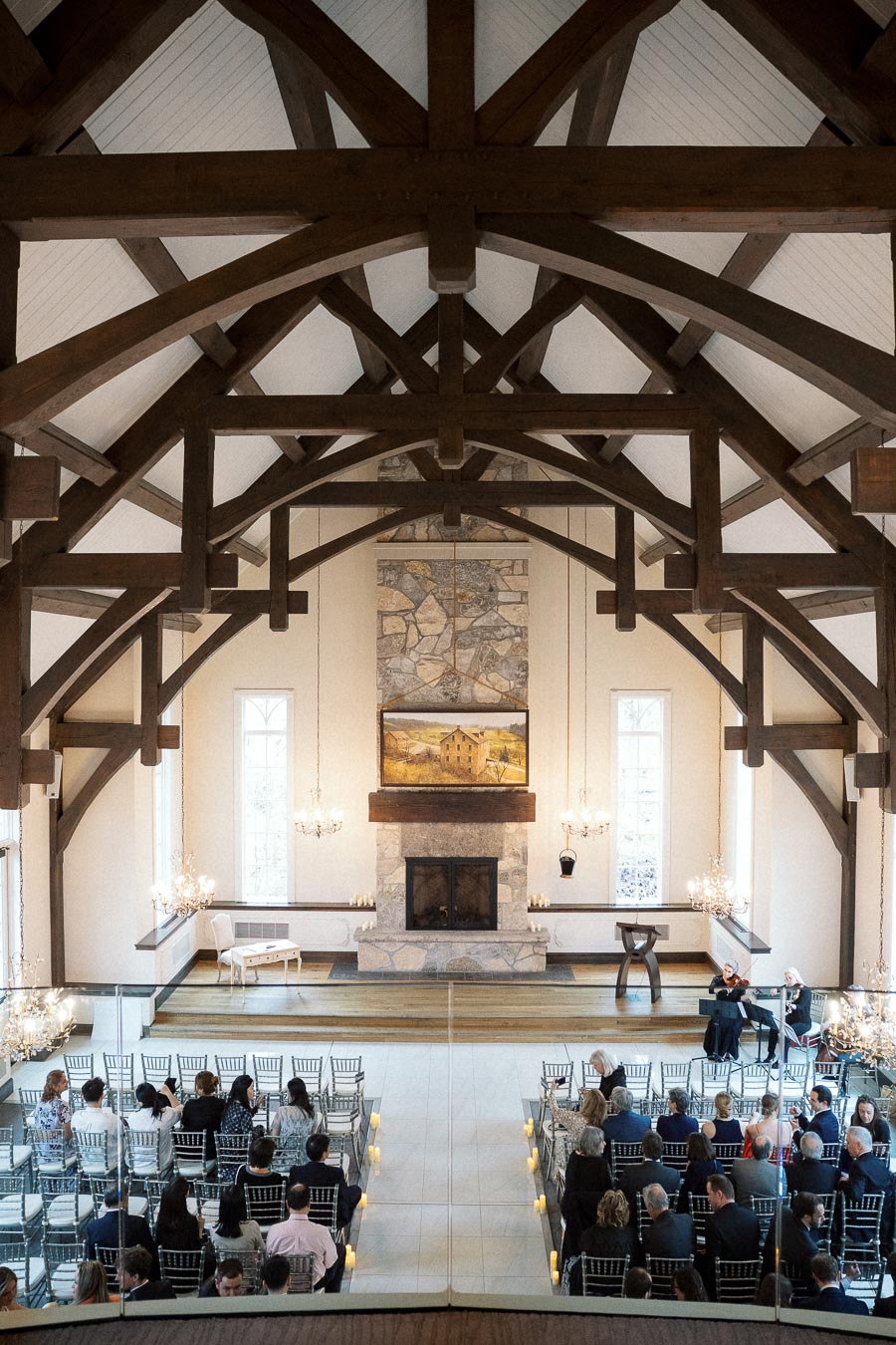Indoor ceremony space with timber beams, fireplace, rows of seated guests, and musicians.