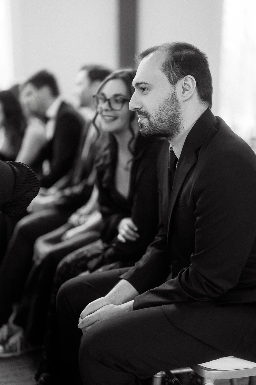 Guests seated indoors, with a man in the foreground and a woman smiling behind him.