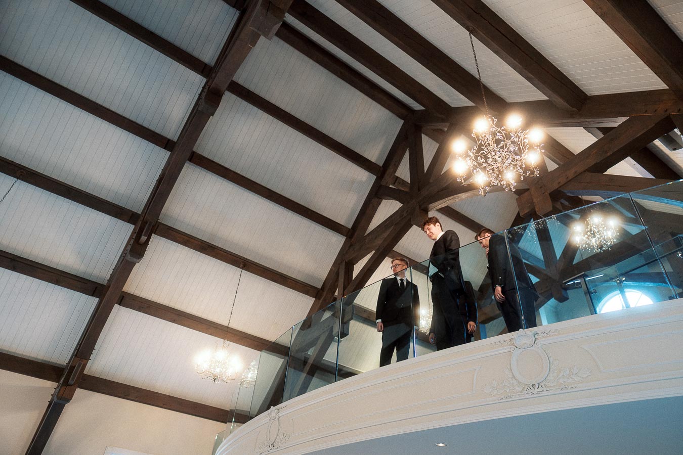 Three men in black suits stand on a glass balcony beneath chandeliers.