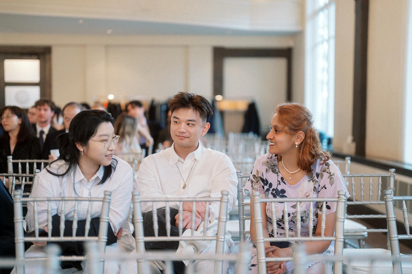 Three guests sitting among silver chairs and talking at an indoor event.
