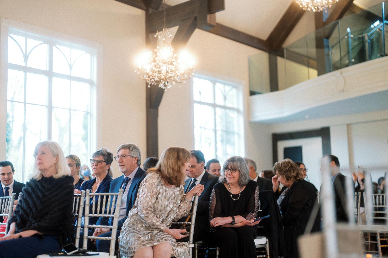 Guests seated indoors beneath chandeliers before a formal ceremony or event.