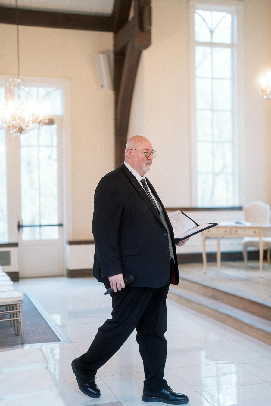 Officiant in a black suit walking indoors with a binder and microphone
