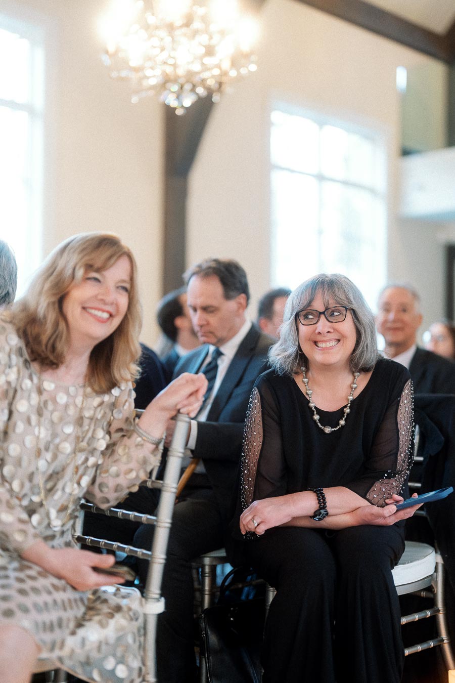 Two seated women smiling during an indoor celebration with guests behind them
