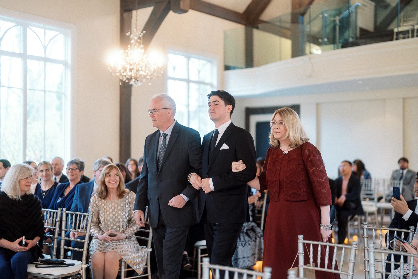 Formally dressed man walks down a ceremony aisle with two older adults.