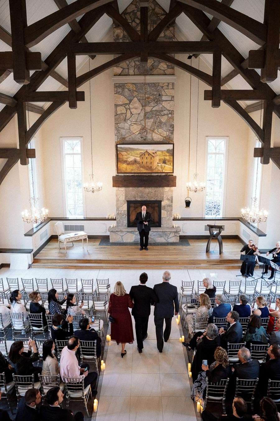 Three people walk down a candle-lined ceremony aisle in a chapel.