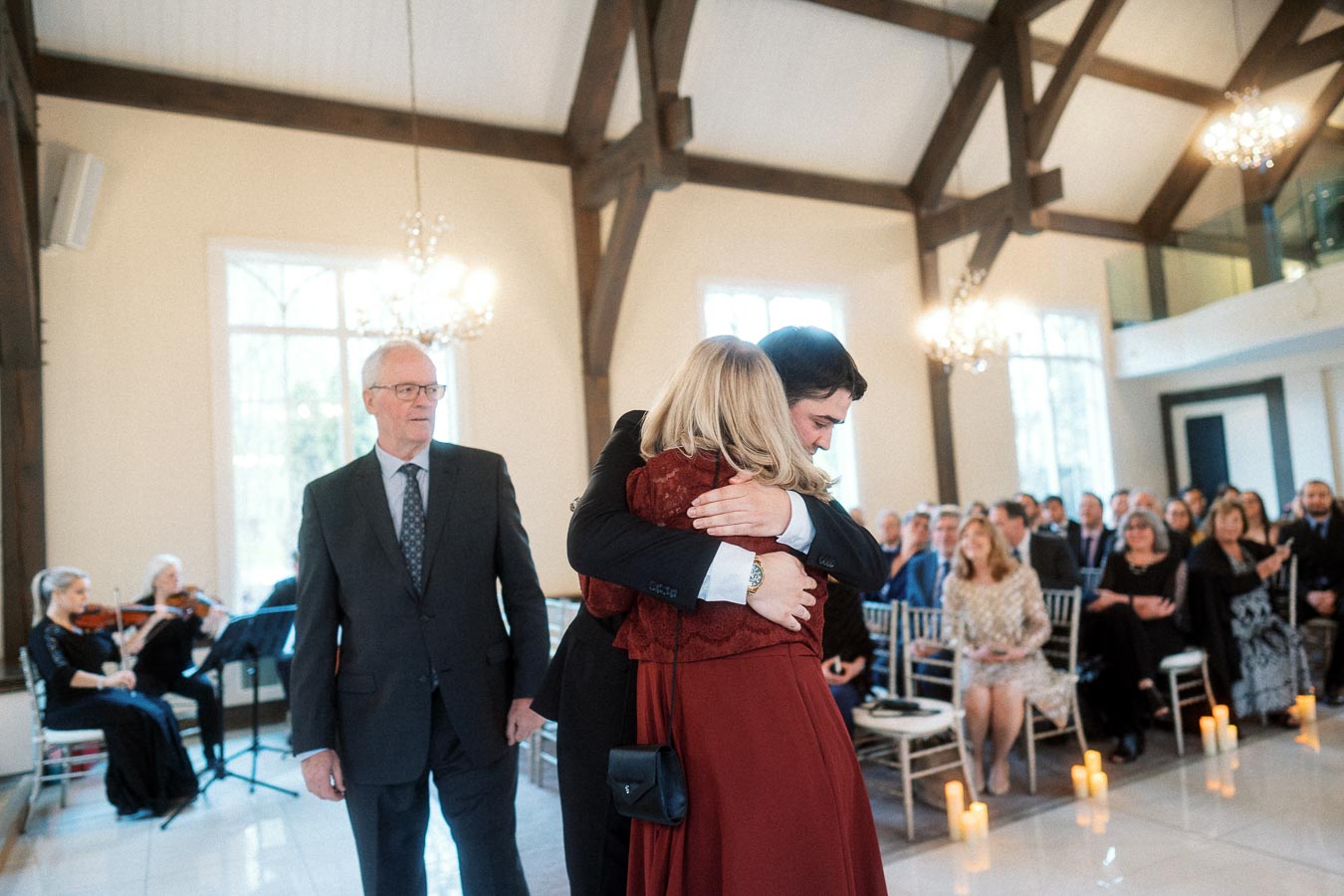 Man hugs woman during an indoor ceremony with seated guests and musicians nearby.