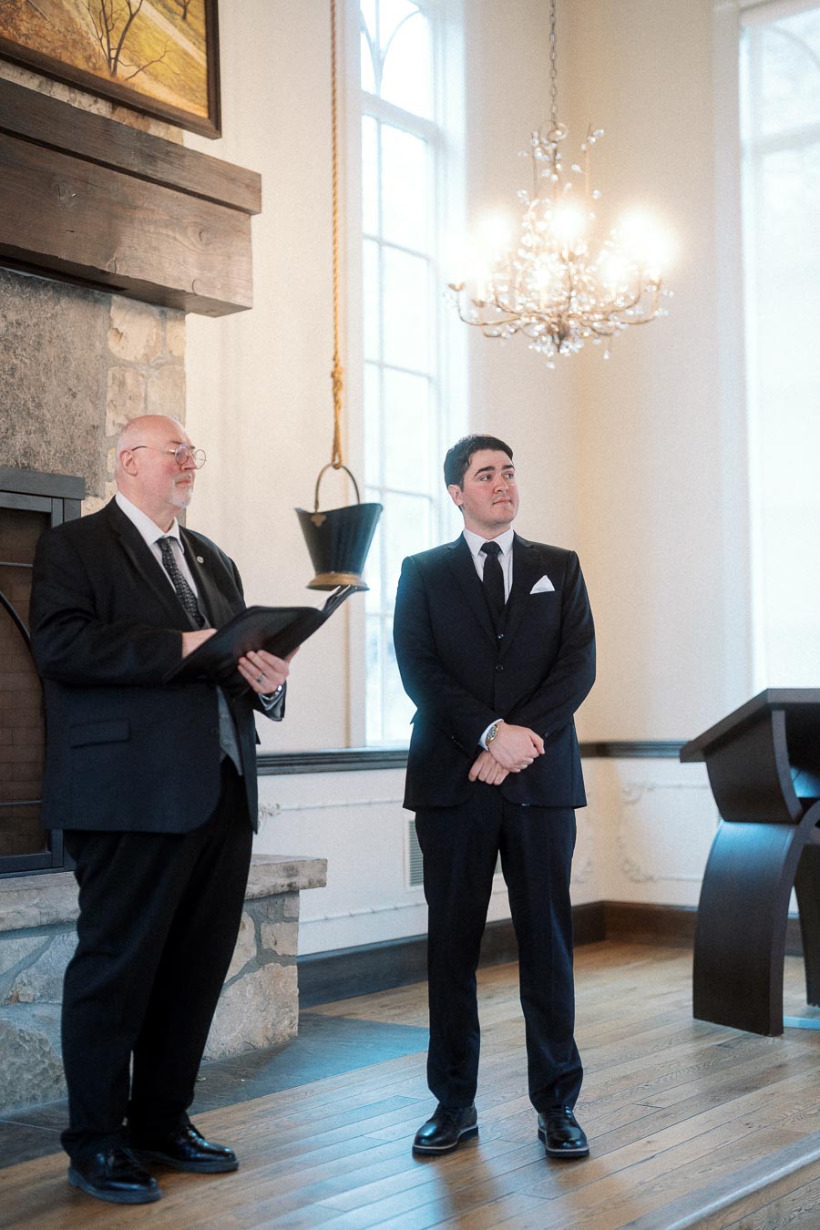 Man in a black suit stands beside an officiant in a bright ceremony room.