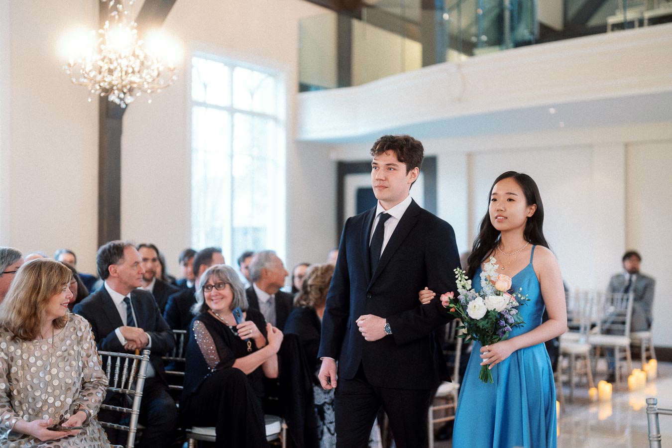 Attendants walk arm in arm down a bright indoor ceremony aisle.