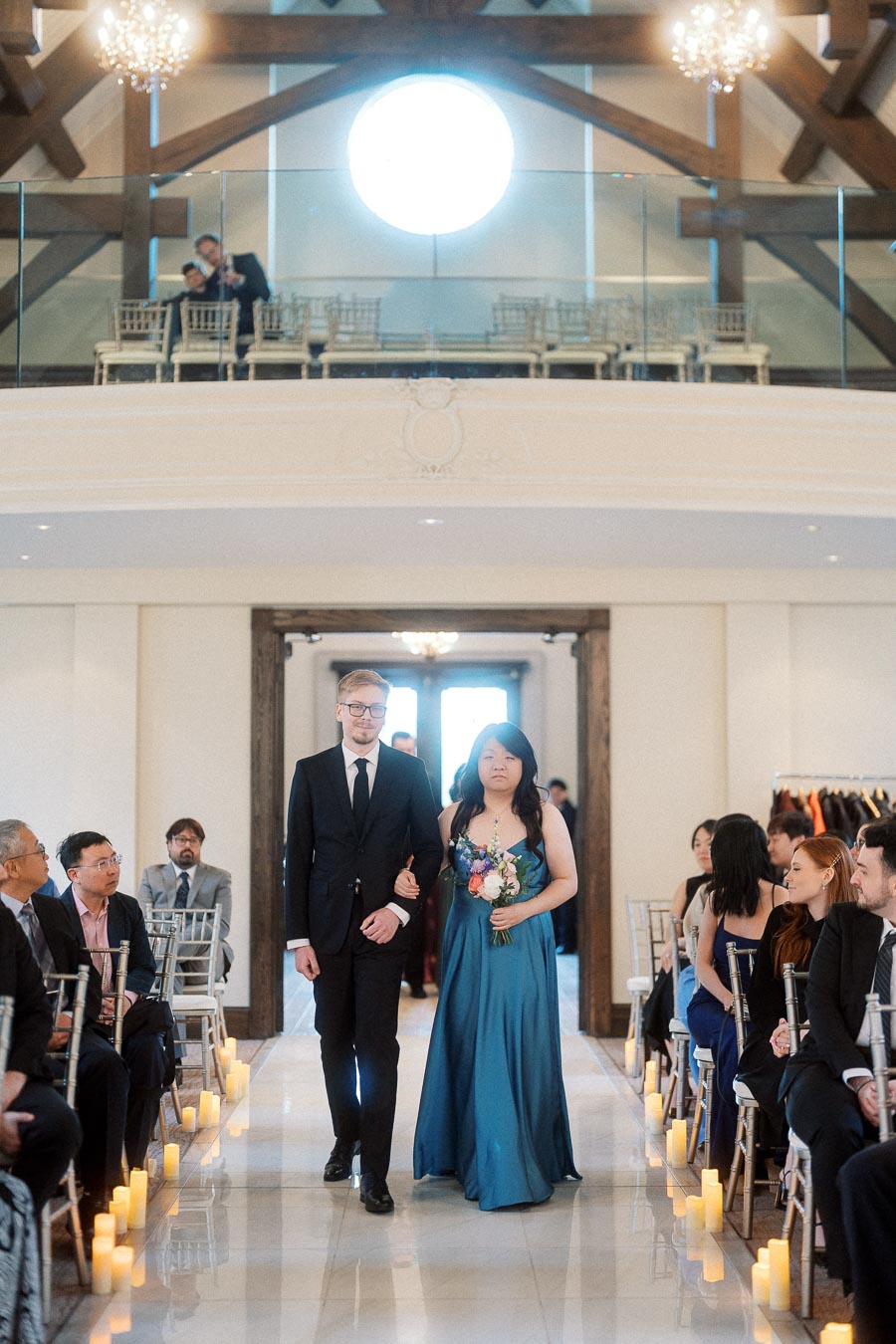 Two formally dressed attendants walk down a candle-lined ceremony aisle with guests seated nearby.
