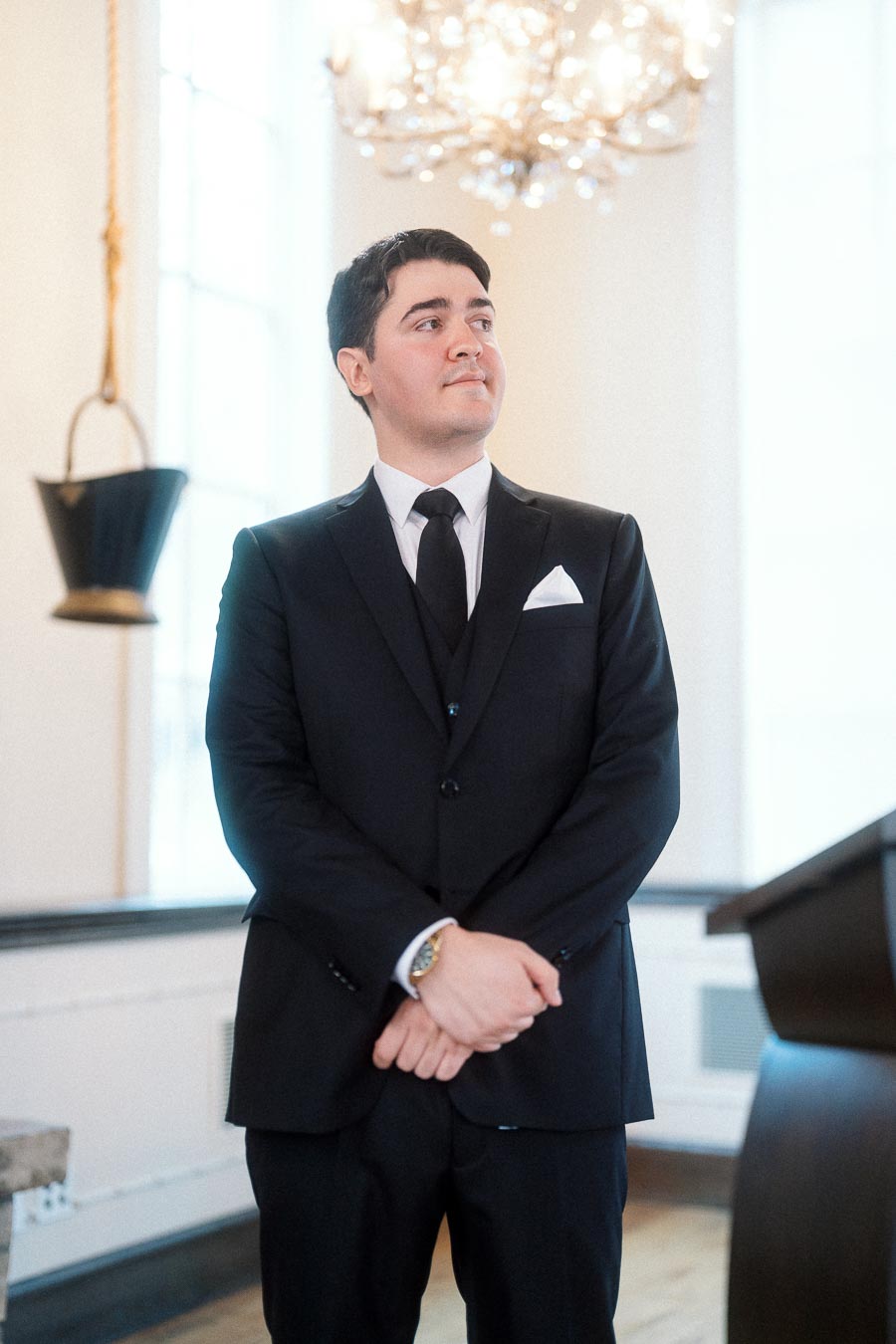 Man in a black suit standing indoors beneath a chandelier