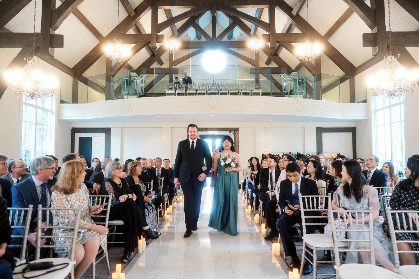 Wedding attendants walk down the aisle in a bright hall filled with seated guests.