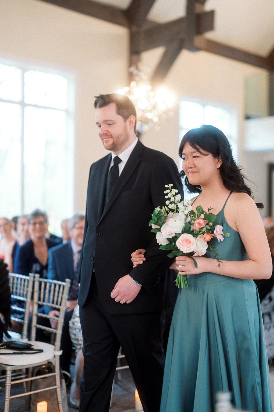 Formally dressed pair walking arm in arm, one holding a bouquet, in an indoor ceremony space