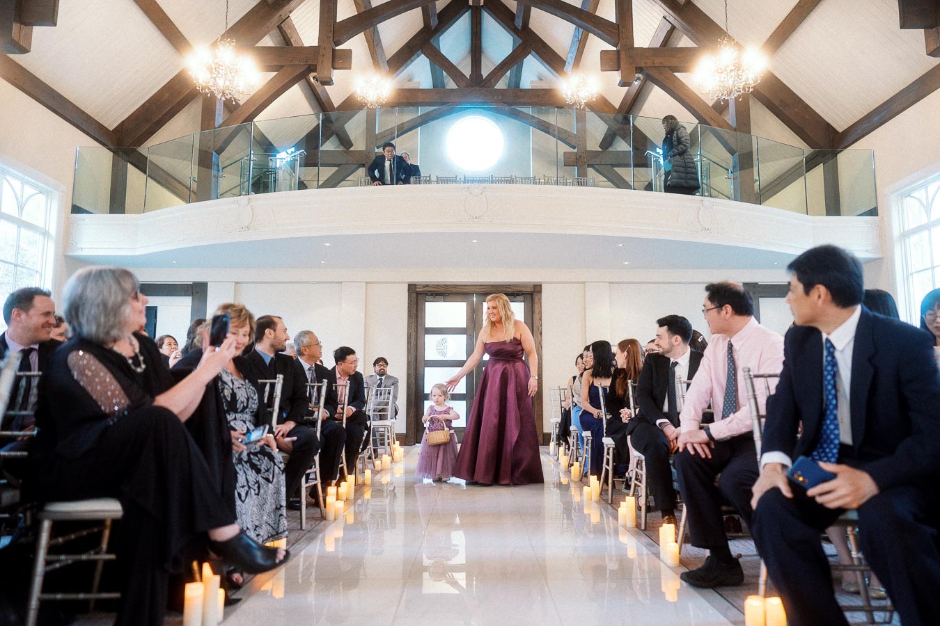 Woman in a burgundy gown and young child walk down a candlelit ceremony aisle.