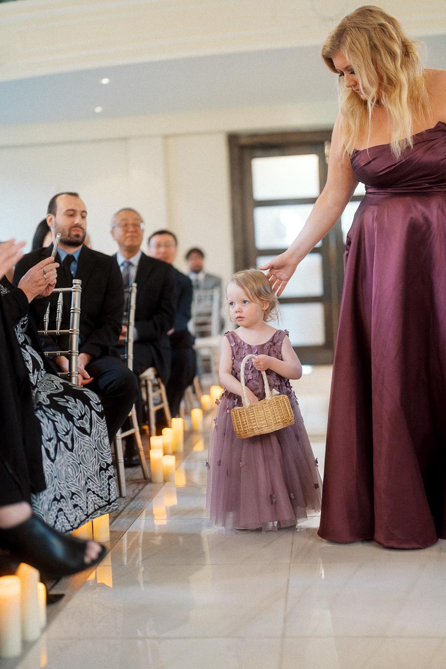 Young flower girl holding a basket beside a woman in a plum dress during an indoor ceremony