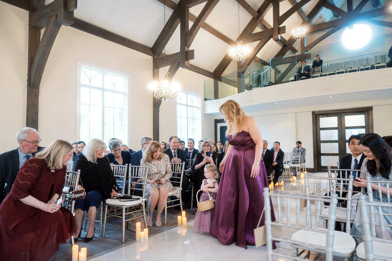 Young flower girl walks down an indoor ceremony aisle beside a woman in a purple gown.
