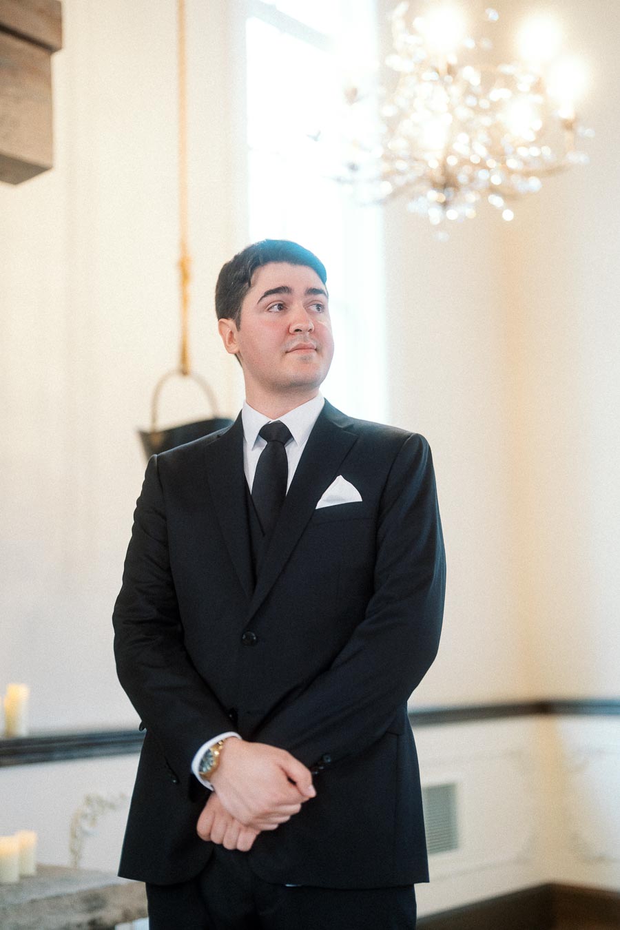 Man in a black suit standing indoors beneath a chandelier