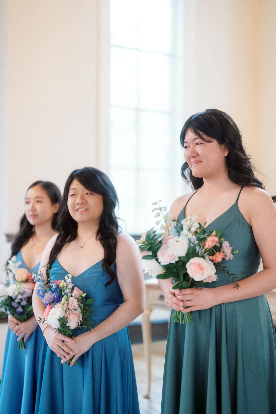 Three women in blue and green dresses holding floral bouquets indoors