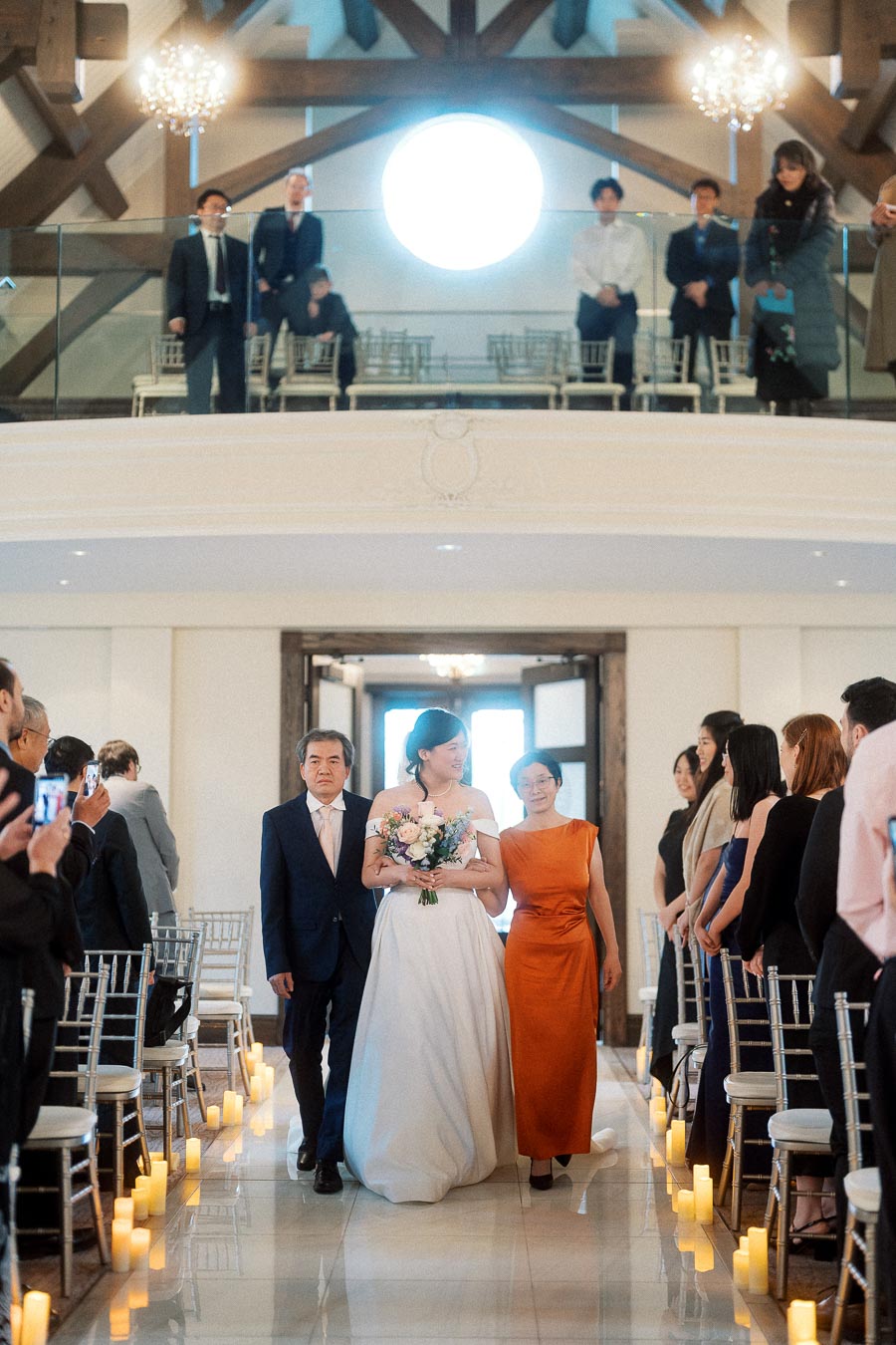 Bride walks down a candlelit indoor aisle with two family members.