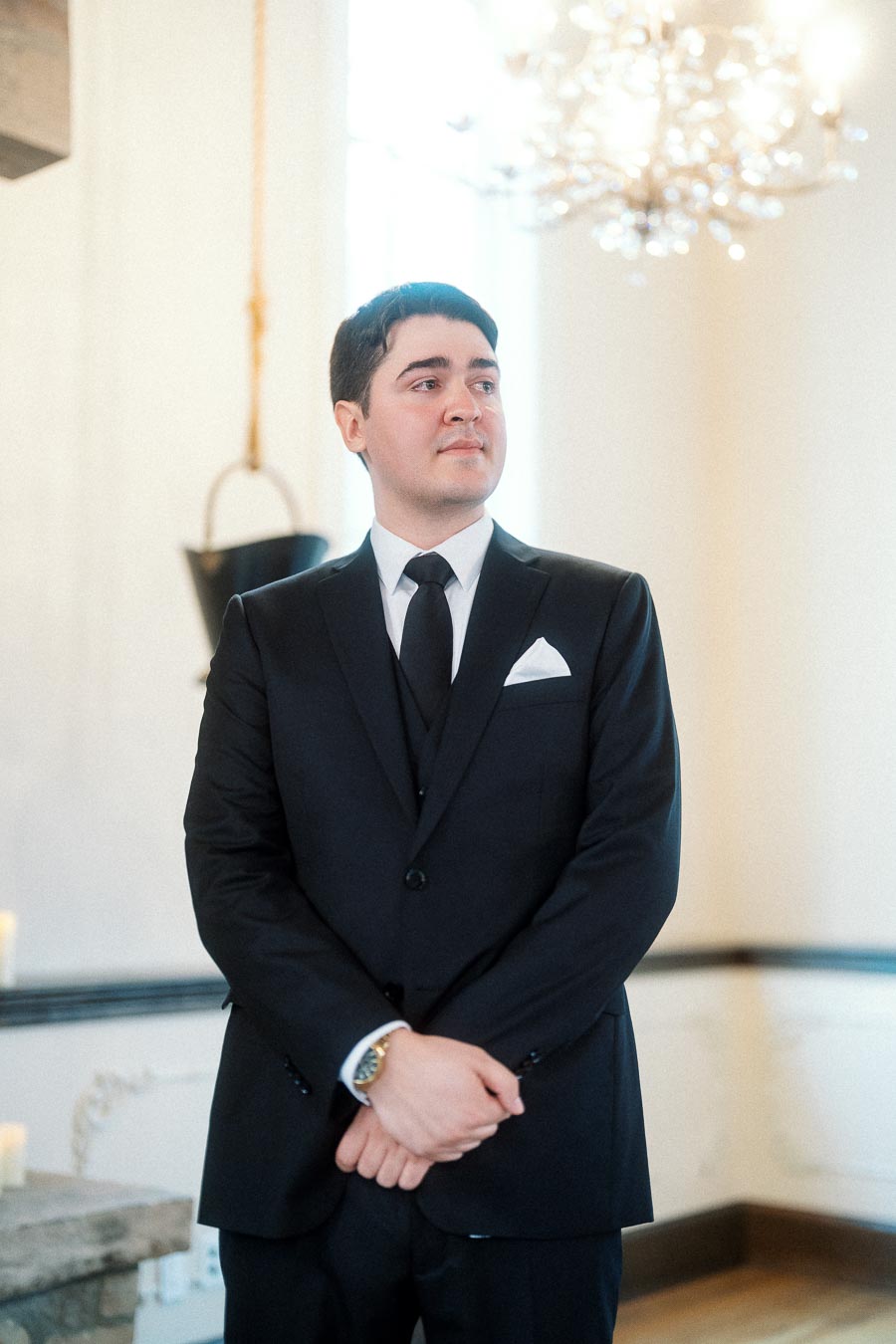 Man in a black suit standing indoors beneath a chandelier.